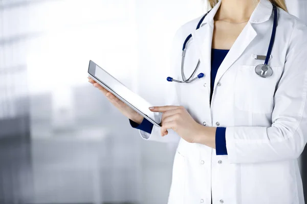 Unknown woman-doctor is holding a tablet computer in her hands, while standing in a clinic. Female physician with a stethoscope in her office, close-up. Perfect medical service in a hospital. Medicine concept.