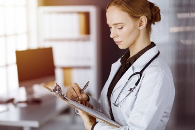 Neşeli gülümseyen kadın doktor güneşli klinikte pano kullanıyor. İş yerindeki dost canlısı doktor kadın portresi. Tıp konsepti