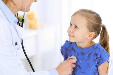 Doktor steteskopla küçük bir kızı muayene ediyor. Her zamanki tıbbi muayenede gülümseyen çocuk hasta. Tıp ve sağlık hizmetleri kavramları