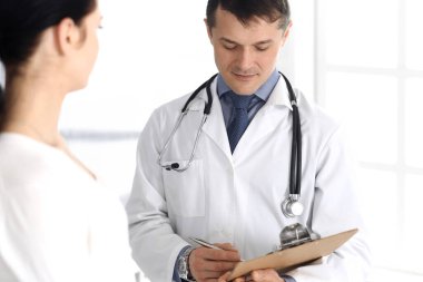 Doktor ve hasta hastane ofisinde tıbbi muayene sonuçlarını tartışıyorlar. Doktor ilaç kayıtlarını doldurmak için pano kullanıyor. Klinikte mükemmel bir tıbbi hizmet. İlaç ve sağlık hizmetleri