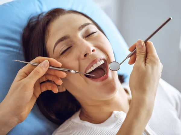 Uśmiechnięta brunetka badana przez dentystę w klinice dentystycznej. Ręce lekarza trzymającego instrumenty stomatologiczne w pobliżu ust pacjenta. Zdrowe zęby i koncepcja medycyny — Zdjęcie stockowe