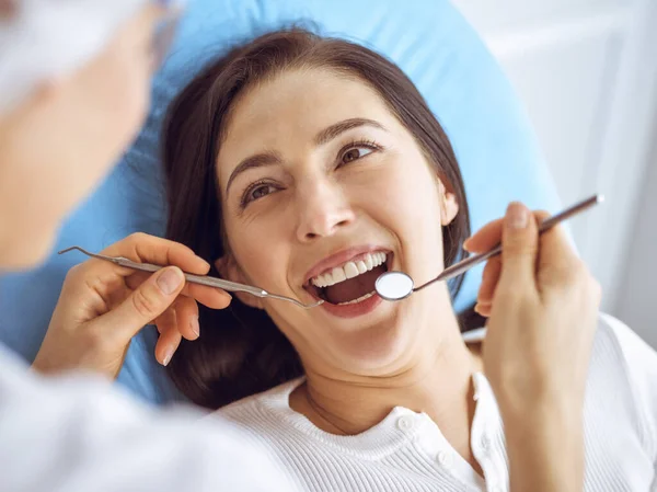 Lachende brunette vrouw wordt onderzocht door tandarts in de tandheelkundige kliniek. Handen van een arts met tandheelkundige instrumenten in de buurt van de mond van patiënten. Gezonde tanden en geneeskundeconcept — Stockfoto