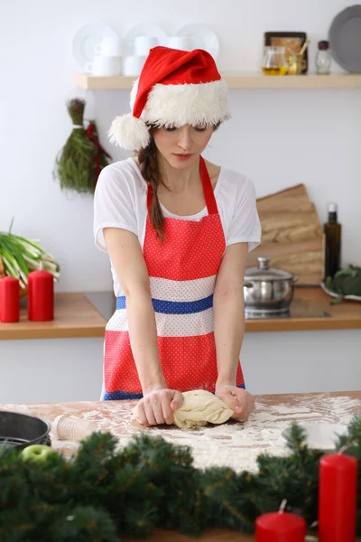 젊은 갈색 머리 여자 가 부엌에서 산타 클로스 모자를 쓰고 피자나 손으로 만든 파스타를 요리한다. 나무 식탁에서 반죽을 준비하는 주부 — 스톡 사진