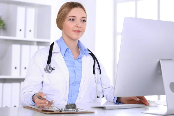 Mujer joven médico en el trabajo en el hospital mirando monitor de escritorio PC. El médico controla los registros de la historia de la medicación y los resultados del examen. Concepto de medicina y salud — Foto de Stock
