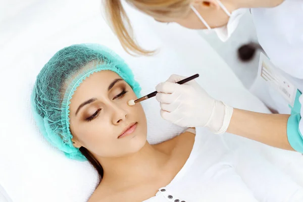 Professionele schoonheidsspecialiste doet cosmetische ingreep op lichte medische achtergrond raken patiënten gezicht met borstel, close-up. Cosmetologische behandeling — Stockfoto