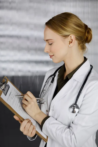 Friendly smiling female doctor using clipboard in clinic. Portrait of friendly physician woman at work. Medical service in hospital. Medicine concept