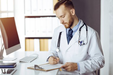 Dost canlısı kızıl sakallı doktor güneşli klinikteki panoda oturuyor ve yazıyor. Tıp konsepti