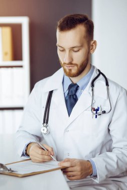 Dost canlısı kızıl sakallı doktor güneşli klinikteki panoda oturuyor ve yazıyor. Tıp konsepti