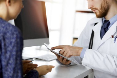 Bilinmeyen kızıl sakallı doktor ve hasta kadın güncel sağlık muayenesini güneşli klinikte otururken ve tablet bilgisayar kullanırken tartışıyorlar, yakın plan. Tıp konsepti