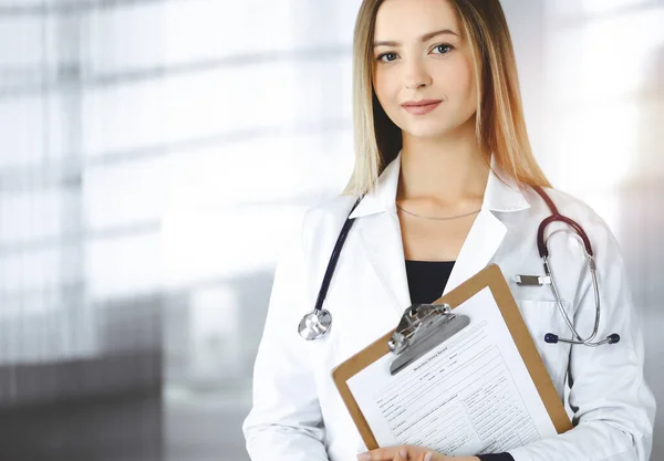 Stock image Young optimistic woman-doctor is holding a clipboard in her hands, while standing in a sunny clinic. Portrait of friendly female physician with a stethoscope. Perfect medical service in a hospital