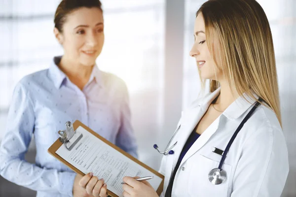 La joven doctora y su paciente están discutiendo el examen de salud actual de los pacientes, mientras están de pie juntos en una soleada oficina del hospital. Médico femenino está escribiendo algunas marcas, utilizando un portapapeles —  Fotos de Stock