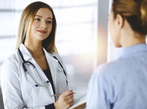 La jeune femme-médecin et sa patiente discutent de l'examen de santé actuel des patients, tout en restant ensemble dans un bureau d'hôpital ensoleillé. Médecin féminin écrit quelques marques, en utilisant un presse-papiers — Photo