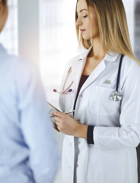 La joven doctora y su paciente están discutiendo el examen de salud actual de los pacientes, mientras están de pie juntos en una soleada oficina del hospital. Médico femenino está escribiendo algunas marcas, utilizando un portapapeles —  Fotos de Stock