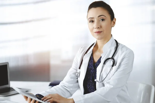 Allegro professionista donna-medico sta utilizzando un computer tablet, mentre seduto nel suo gabinetto soleggiato in una clinica. Ritratto di medico donna amichevole con stetoscopio al lavoro. Servizio medico perfetto — Foto Stock