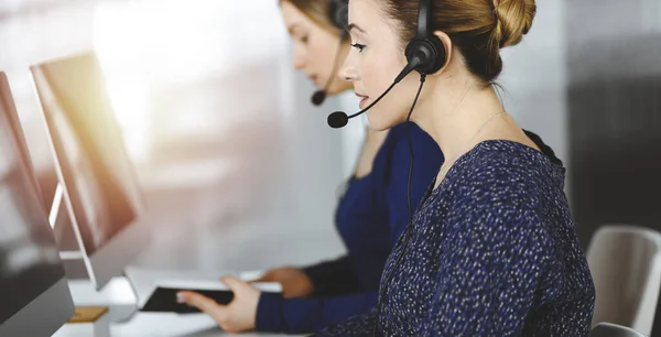 Two busineswomen have conversations with the clients by headsets, while sitting at the desk in a sunny modern office. Diverse people group in a call center