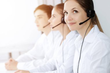 Merkezi ara. Güzel genç bir kadın güneşli ofisteki müşterilere yardım etmek için kulaklık ve bilgisayar kullanıyor. İş konsepti