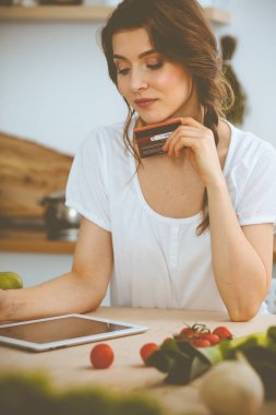 Genç bir kadın mutfakta yemek pişirmek için yeni bir tarif arıyor. Ev hanımı tablet bilgisayar ve kredi kartıyla online alışveriş yapıyor.