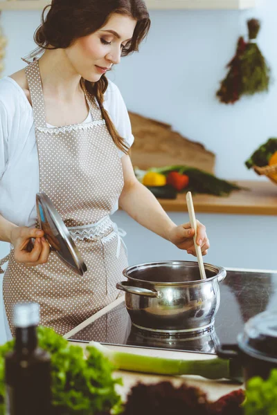 Młoda brunetka gotuje zupę w kuchni. Gospodyni domowa trzymająca drewnianą łyżkę w ręku. Koncepcja żywności i zdrowia — Zdjęcie stockowe