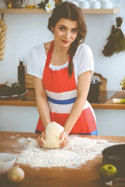 Νεαρή μελαχρινή γυναίκα μαγειρεύει πίτσα ή χειροποίητα ζυμαρικά στην κουζίνα. Νοικοκυρά ετοιμάζει ζύμη σε ξύλινο τραπέζι. Έννοια της διατροφής, των τροφίμων και της υγείας — Φωτογραφία Αρχείου