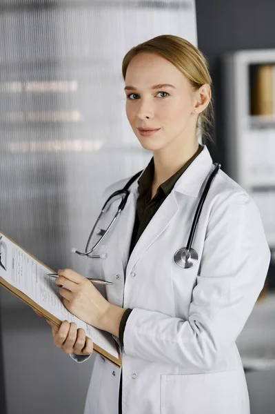 Friendly smiling female doctor using clipboard in clinic. Portrait of friendly physician woman at work. Medical service in hospital. Medicine concept