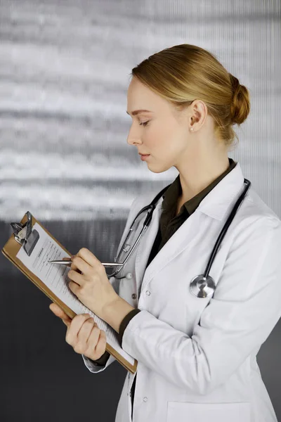 Friendly smiling female doctor using clipboard in clinic. Portrait of friendly physician woman at work. Medical service in hospital. Medicine concept