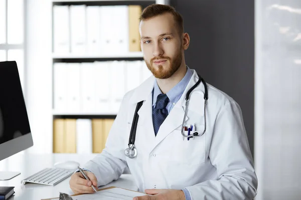 Vriendelijke rode baard dokter zittend en schrijvend aan klembord in kliniek. Perfecte medische dienst in het ziekenhuis. Geneesmiddelenconcept — Stockfoto