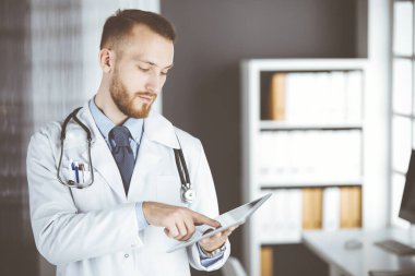Dost canlısı kızıl sakallı doktor çalıştığı yerdeki klinikte tablet bilgisayar kullanıyor. Hastane ve tıp konseptinde mükemmel bir tıbbi hizmet.