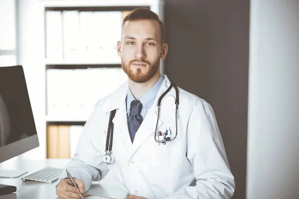 Přátelský rudovousý doktor sedící a píšící na poličce na klinice. Perfektní lékařská služba v nemocnici. Koncept medicíny — Stock fotografie