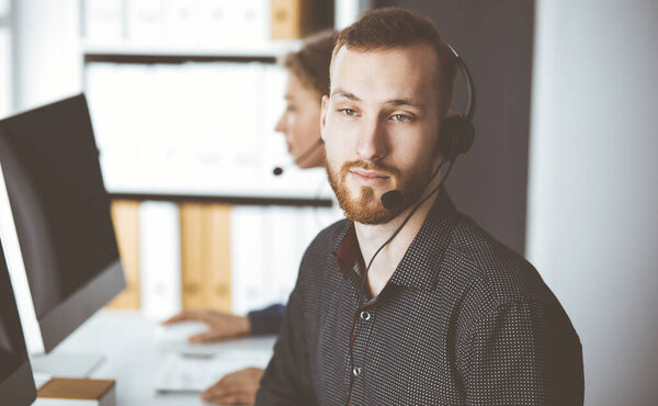 Red-bearded businessman talking by headset near his female colleague while sitting in modern office. Diverse people group in call center. Telemarketing and customer service