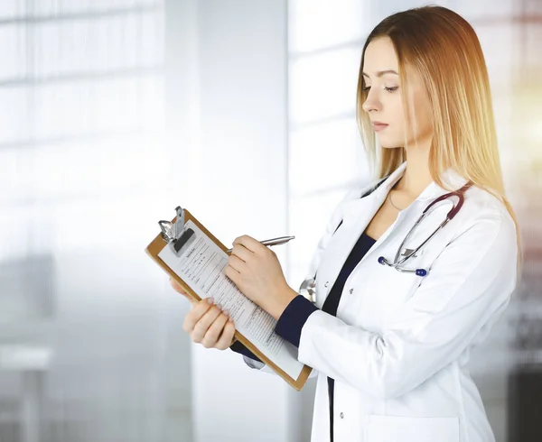 Jovem mulher-médico otimista está segurando uma prancheta em suas mãos, enquanto está em uma clínica ensolarada. Retrato de médico feminino amigável com um estetoscópio. Serviço médico perfeito em um hospital — Fotografia de Stock