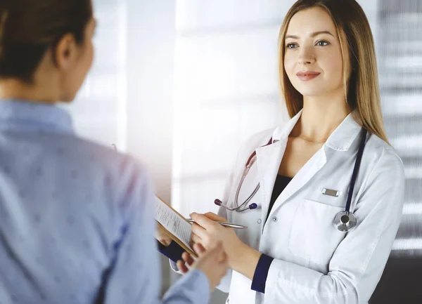 La jeune femme-médecin et sa patiente discutent de l'examen de santé actuel des patients, tout en restant ensemble dans un bureau d'hôpital ensoleillé. Médecin féminin écrit quelques marques, en utilisant un presse-papiers — Photo