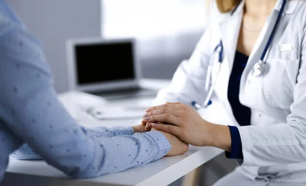 La desconocida doctora está sosteniendo las manos de sus pacientes, discutiendo el examen de salud actual, mientras están sentados en el escritorio en el gabinete de una clínica. Médico con un estetoscopio en el trabajo, primer plano — Foto de Stock