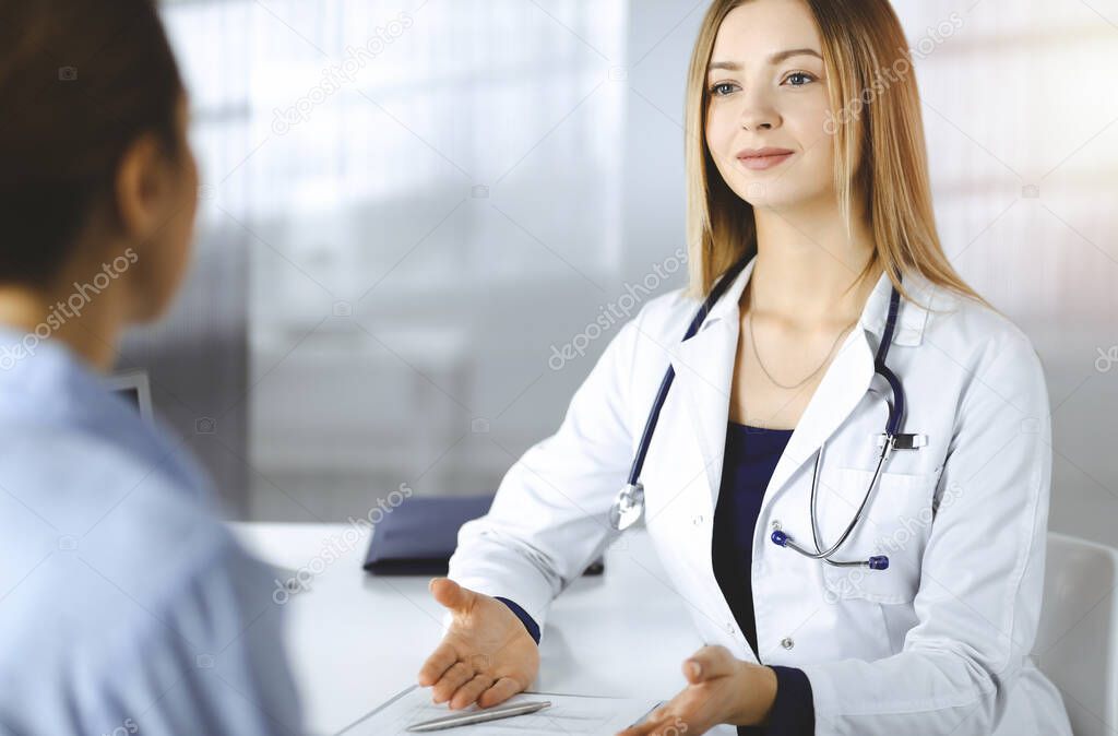 Unknown woman-doctor is writing some medical recommendations to her patient, while they are sitting together at the desk in the sunny cabinet in a clinic. Physician is using a clipboard, close-up
