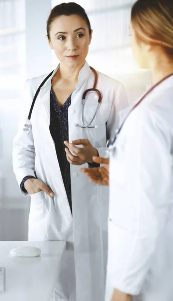 Two female physicians are talking about medical therapy, while standing together in a sunny clinic office. Doctors use stethoscopes at work. Teamwork in medicine