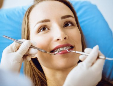 Diş hekiminde dişçi tarafından muayene edilen diş telleriyle gülümseyen genç bir kadın. Sağlıklı dişler ve tıbbi bakım konsepti