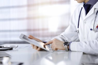 Bilinmeyen erkek doktor, tablet bilgisayarla oturuyor ve çalışıyor. Karanlık bir klinikte, arka planda ışık parıltısı, ellerin yakın çekimi.