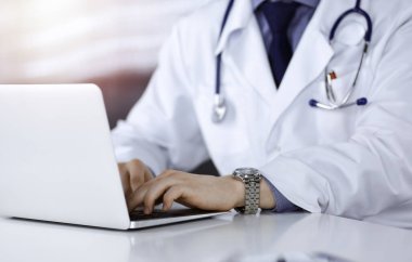 Bilinmeyen bir erkek doktor karanlık bir klinikte dizüstü bilgisayarla oturuyor ve çalışıyor. Arka planda ışık parıltısı var.