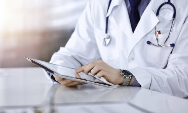 Bilinmeyen erkek doktor, tablet bilgisayarla oturuyor ve çalışıyor. Karanlık bir klinikte, arka planda ışık parıltısı, ellerin yakın çekimi.