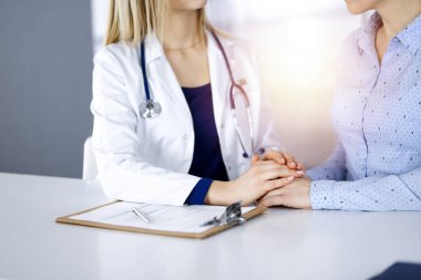 Bilinmeyen kadın doktor, bir klinikteki güneşli dolapta yan yana otururken hastasına güven vermek için elini tutuyor. Kadın hekim bir pano ve steteskop kullanıyor.