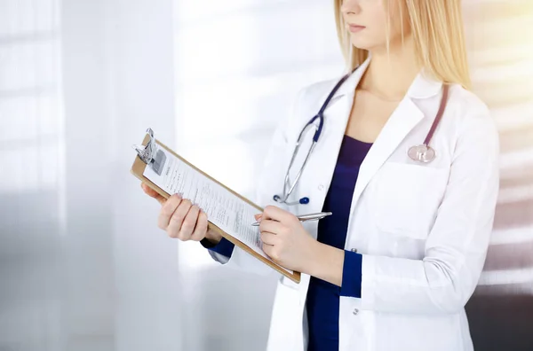 신원을 알 수없는 여성 - 의사는 클리닉에서 햇볕 이 잘 드는 캐비닛에 서 있는 동안 클립보드를 사용 해 몇 가지 노트를 쓰고 있다. 청진기를 가지고 있는 여성 의사, 클로즈업. 병원에서의 완벽 한 의료 서비스 — 스톡 사진