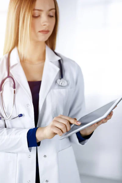 젊은 전문 여성 과 의사가 햇빛 이 잘 드는 병원에 서 있는 동안 태블릿 컴퓨터를 사용하고 있습니다. 아름다운 여성 의사가 활동하는 모습. 병원에서의 완벽 한 의료 서비스. 의학적 개념 — 스톡 사진