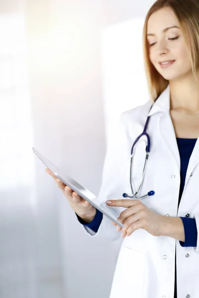 Mladá žena-doktorka drží v rukou tablet, zatímco stojí na slunné klinice. Portrét přátelského ženského lékaře se stetoskopem v práci. Perfektní lékařská služba v nemocnici — Stock fotografie