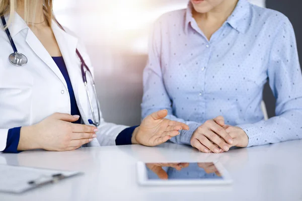 Mulher-médico desconhecido está mostrando a seu paciente uma descrição da medicação, enquanto sentados juntos na mesa no armário ensolarado em uma clínica. Médica feminina está usando um tablet de computador e um — Fotografia de Stock