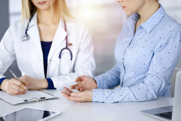 Femme inconnue- médecin écoute sa patiente, alors qu'elle est assise ensemble au bureau dans l'armoire ensoleillée d'une clinique. Une femme médecin avec un stéthoscope écrit au presse-papiers, gros plan. Parfait. — Photo