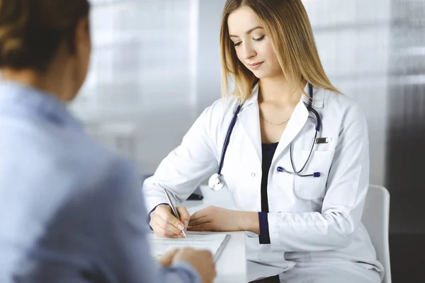 Giovane donna-medico intelligente sta ascoltando i sintomi dei suoi pazienti, mentre si siede insieme nell'armadietto in una clinica. Il medico, con un sorriso, sta scrivendo alcuni appunti sui suoi documenti al — Foto Stock