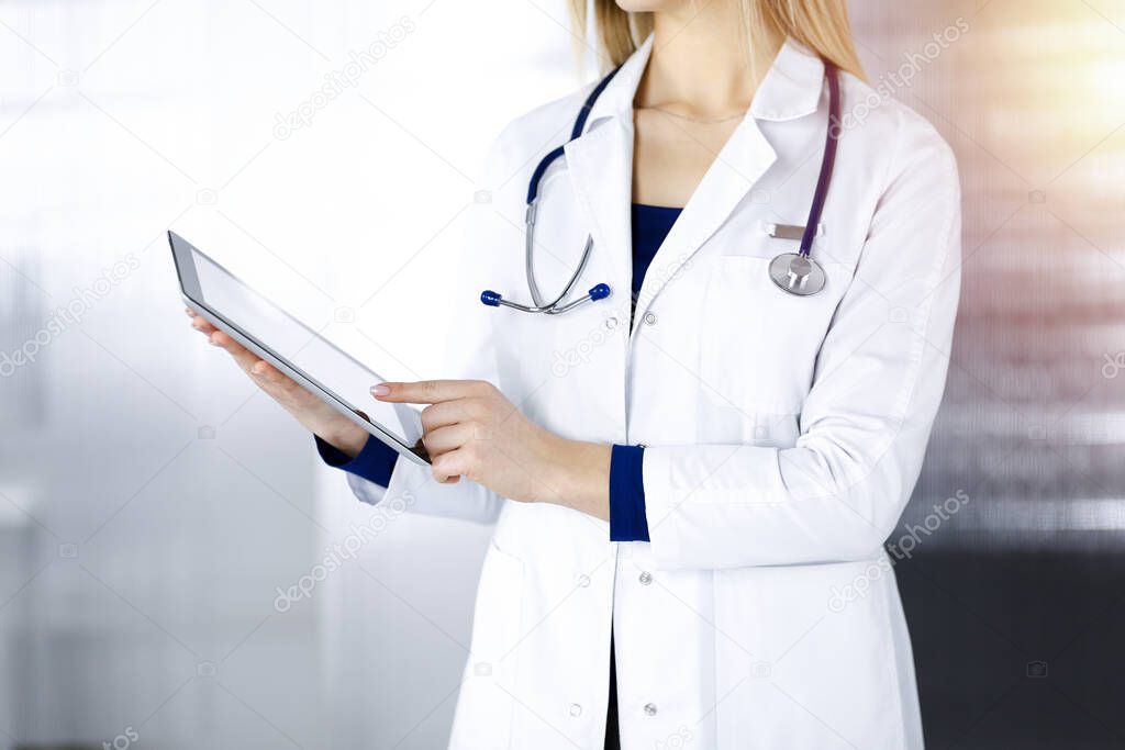 Unknown woman-doctor is holding a tablet computer in her hands, while standing in a sunny clinic cabinet. Female physician at work, close-up. Perfect medical service in a hospital. Medicine concept