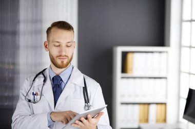 Dost canlısı kızıl sakallı doktor çalıştığı yerdeki klinikte tablet bilgisayar kullanıyor. Hastane ve tıp konseptinde mükemmel bir tıbbi hizmet.