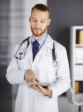 Dost canlısı kızıl sakallı doktor çalıştığı yerdeki klinikte tablet bilgisayar kullanıyor. Hastane ve tıp konseptinde mükemmel bir tıbbi hizmet.