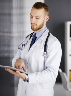 Dost canlısı kızıl sakallı doktor çalıştığı yerdeki klinikte tablet bilgisayar kullanıyor. Hastane ve tıp konseptinde mükemmel bir tıbbi hizmet.