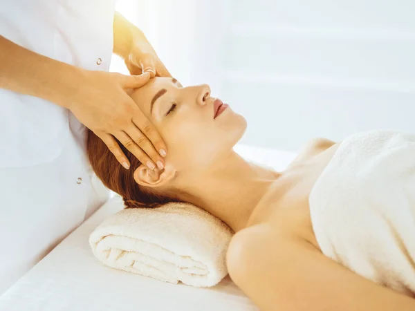Beautiful woman enjoying facial massage with closed eyes in sunny spa center. Relaxing treatment concept in medicine — Stock Photo, Image